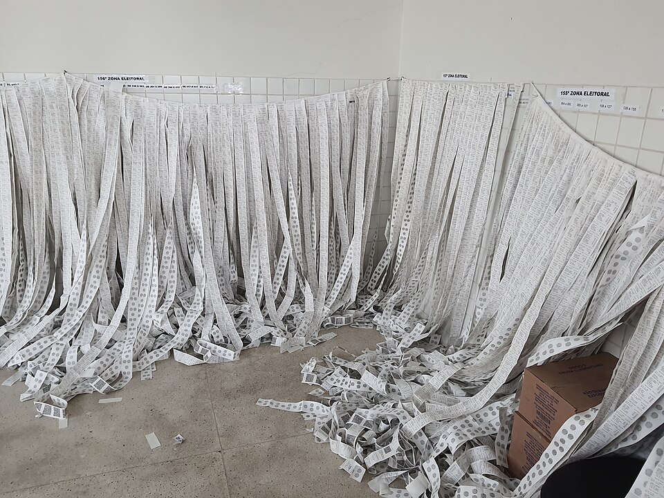 Dezenas de boletins de urna impressos pendurados em varais numa sala de cartório eleitoral