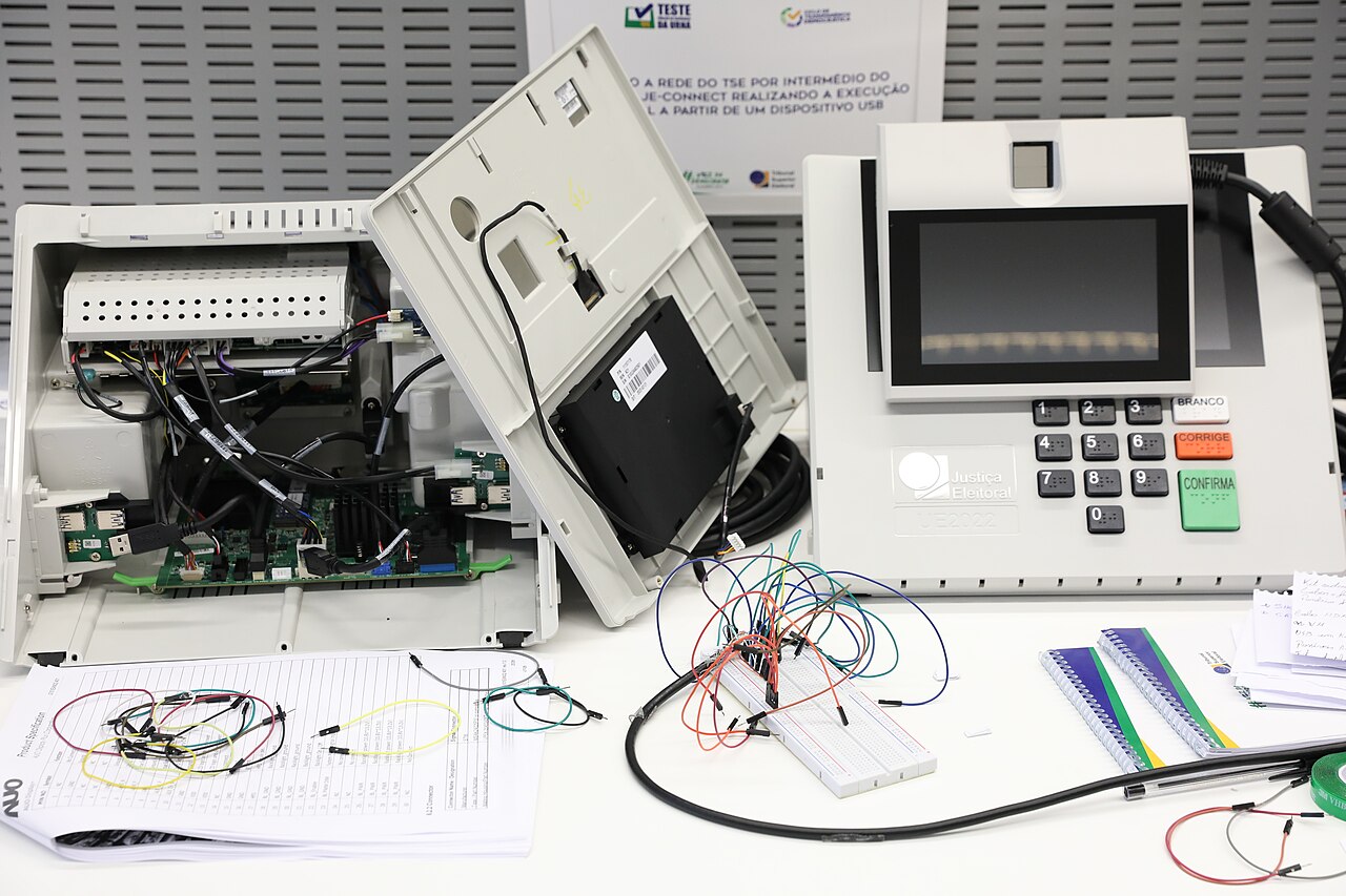 Urna eletrônica aberta sobre uma mesa, com placas e cabos expostos ao lado de outra urna fechada